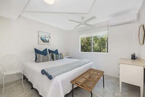 Ein Schlafzimmer mit Bett, Schreibtisch, Stuhl, Deckenventilator und einem Fenster mit Blick auf Bäume.