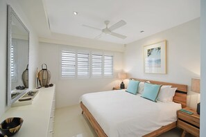 Ein Schlafzimmer mit Bett, Nachttischen, Deckenventilator, Fenster mit Jalousien und einem Wandbild.