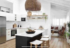 A modern kitchen with white cabinetry, a central island, a hanging light fixture, and a view into a living area with a sofa and chairs. - The Lodge Noosa Coastal Luxe in Noosas Heart (Noosa)