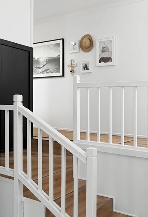 Eine Treppe mit weißen Geländern, eine dunkle Tür sowie eine Wand mit gerahmten Bildern und einem Hut.