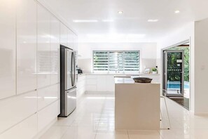 A modern kitchen with a refrigerator, a window with blinds, and a view of a pool. - Sleek Stylish Family Beach House Near Junction (Noosa)