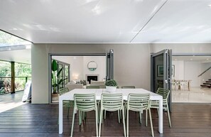 A modern dining area with a white table and green chairs, a fireplace, and a view of the outdoors. - Sleek Stylish Family Beach House Near Junction (Noosa)