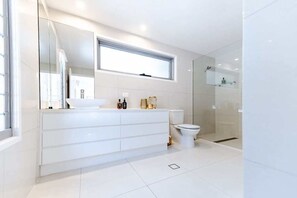 A modern bathroom with a white vanity, a toilet, a shower area, and a large mirror. - Sleek Stylish Family Beach House Near Junction (Noosa)