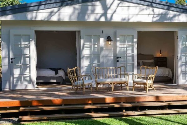 A covered outdoor seating area with wicker furniture, a wooden deck, and a view of a bedroom with a bed and nightstand. - The River Shack Vintage Luxe Right Beside River (Noosa)