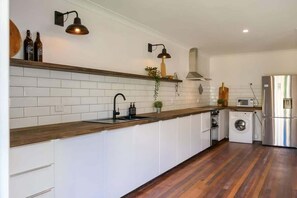 A modern kitchen with white cabinets, a wooden countertop, a stainless steel refrigerator, a washing machine, and two wall-mounted light fixtures. - The River Shack Vintage Luxe Right Beside River (Noosa)