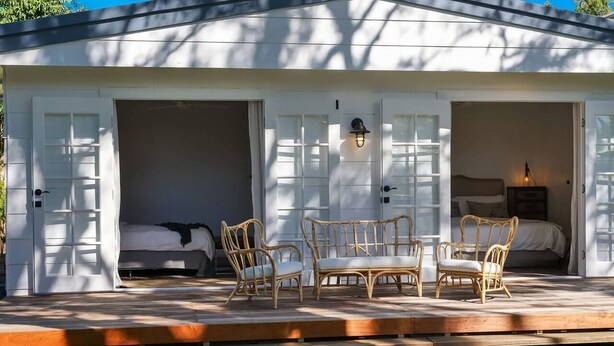 A white building with two open doors leading to bedrooms, each with a bed and a nightstand. There is a wooden deck with wicker furniture.