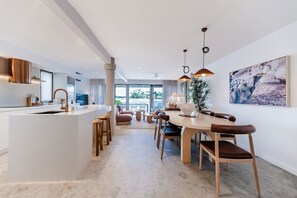 A modern kitchen with a central island, dining area with a wooden table and chairs, and a large window providing a view of the outdoors. - Bella Vista Noosa Luxury on White Sands (Noosa)
