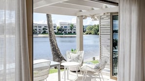 Een moderne kamer met een grote schuifdeur die uitkomt op een terras met een tafel en stoelen, een uitzicht op een waterpartij en een gebouw op de achtergrond.