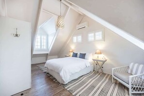 A bedroom with a sloped ceiling, a bed with white bedding, two bedside lamps, a chair, and a patterned rug.