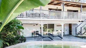 Une piscine avec des chaises longues, une terrasse couverte et un bâtiment à deux étages doté d’un balcon.