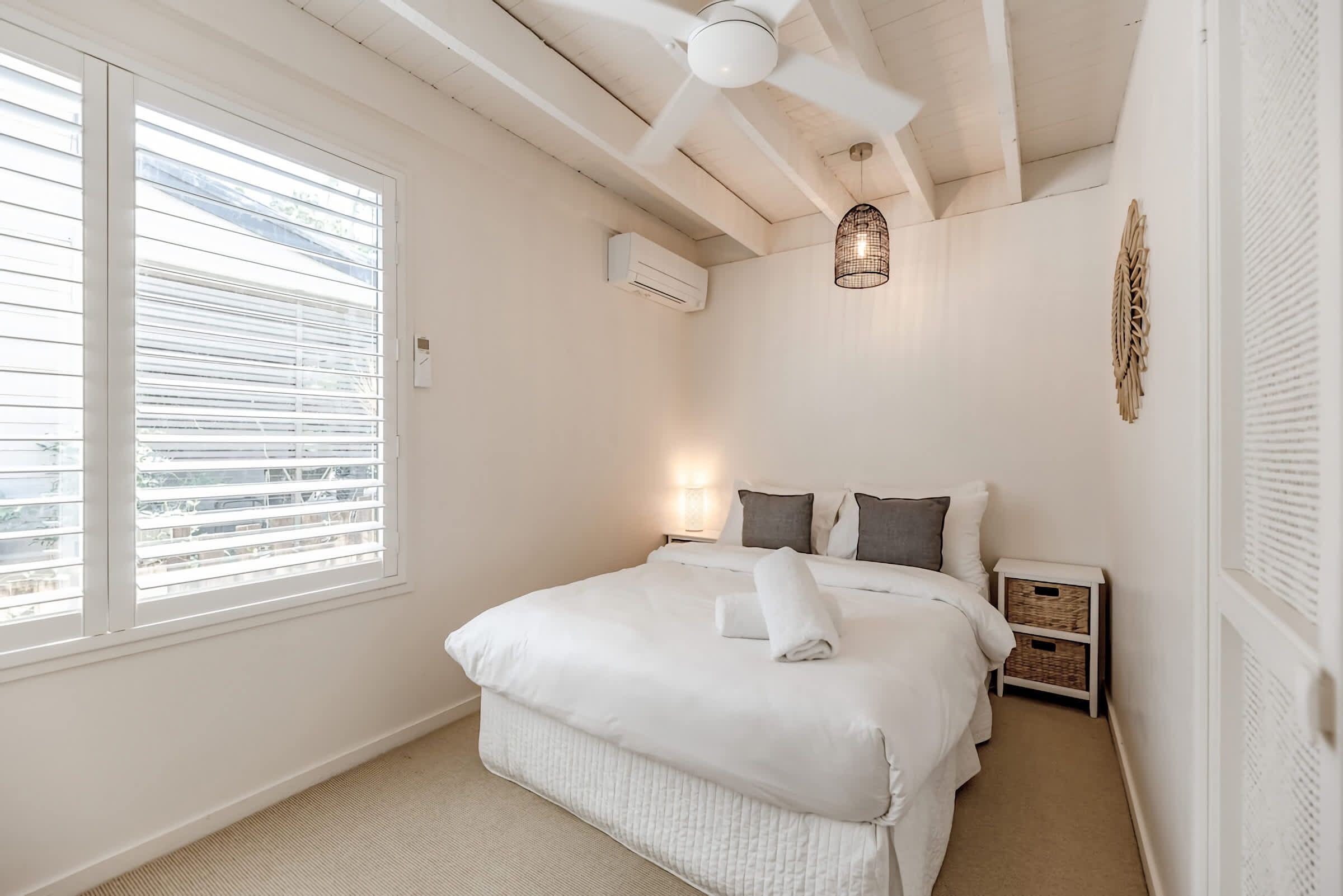 Ein Schlafzimmer mit Bett, Deckenventilator, Klimaanlage, Nachttischlampe und Blick durch das Fenster.