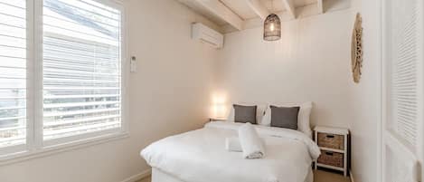 A bedroom with a bed, a ceiling fan, an air conditioning unit, a bedside lamp, and a view through the window.