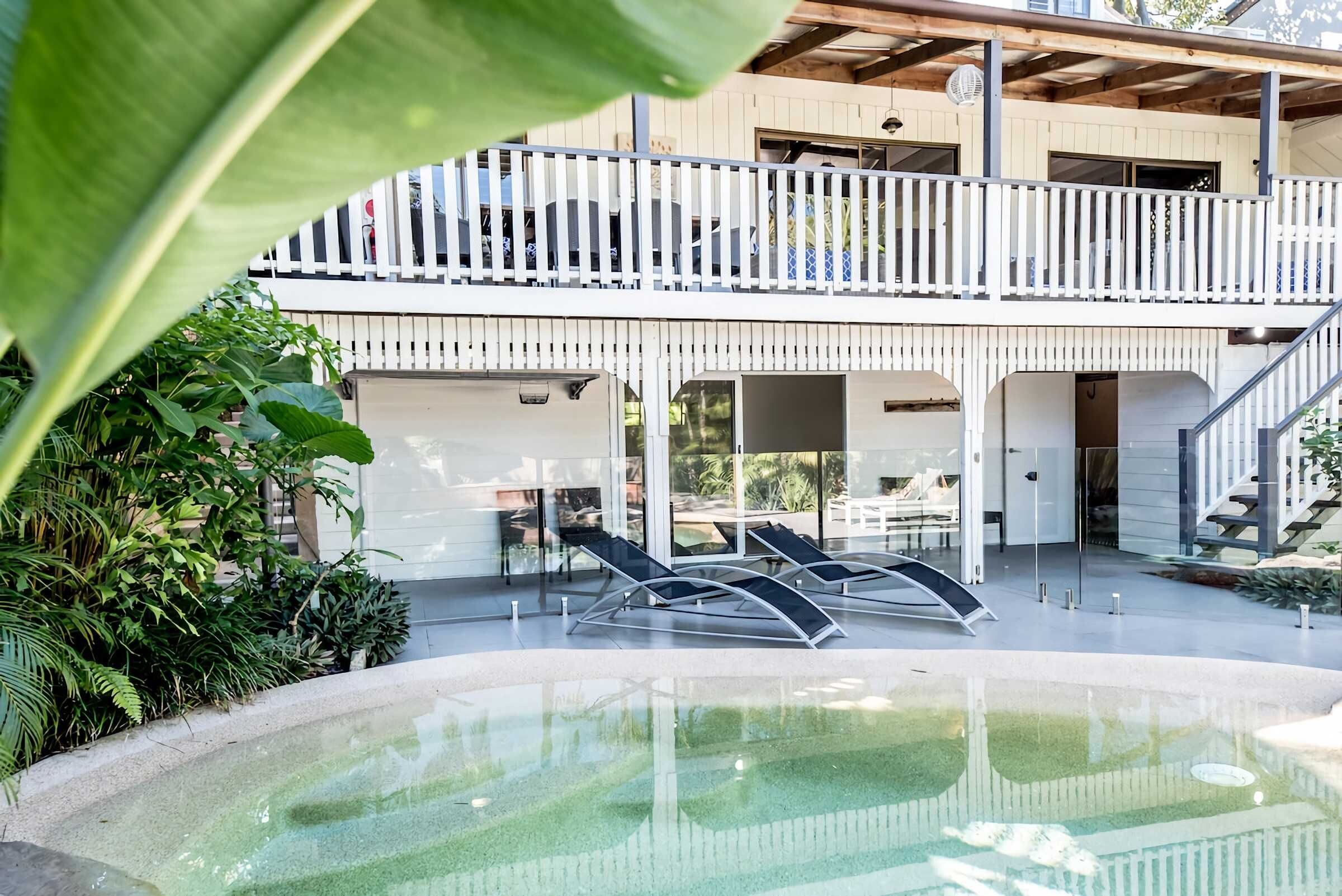 A modern outdoor pool area with lounge chairs, a covered seating area, and a multi-level structure with a balcony.
