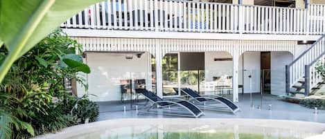 A modern outdoor pool area with lounge chairs, a covered seating area, and a multi-level structure with a balcony.