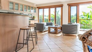 A modern kitchen with wooden cabinets, a bar area with stools, a grey sofa, a wooden coffee table, and large sliding glass doors to a balcony. - Seahorse Family & Pooch Friendly Waterfront Home (Noosa)