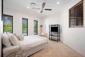 A bedroom with a bed, a ceiling fan, a TV on a black stand, and windows with blinds. - Seahorse Family & Pooch Friendly Waterfront Home (Noosa)