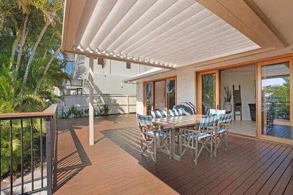 An outdoor deck with a wooden table and chairs, a pergola, and a view of a building with large windows.