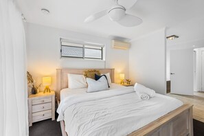 Un dormitorio con cama, mesitas de noche, ventilador de techo, ventana y aire acondicionado.