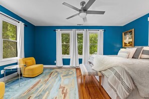 A room with a blue wall, a ceiling fan, a bed with white bedding, a yellow armchair, and a window with white curtains.