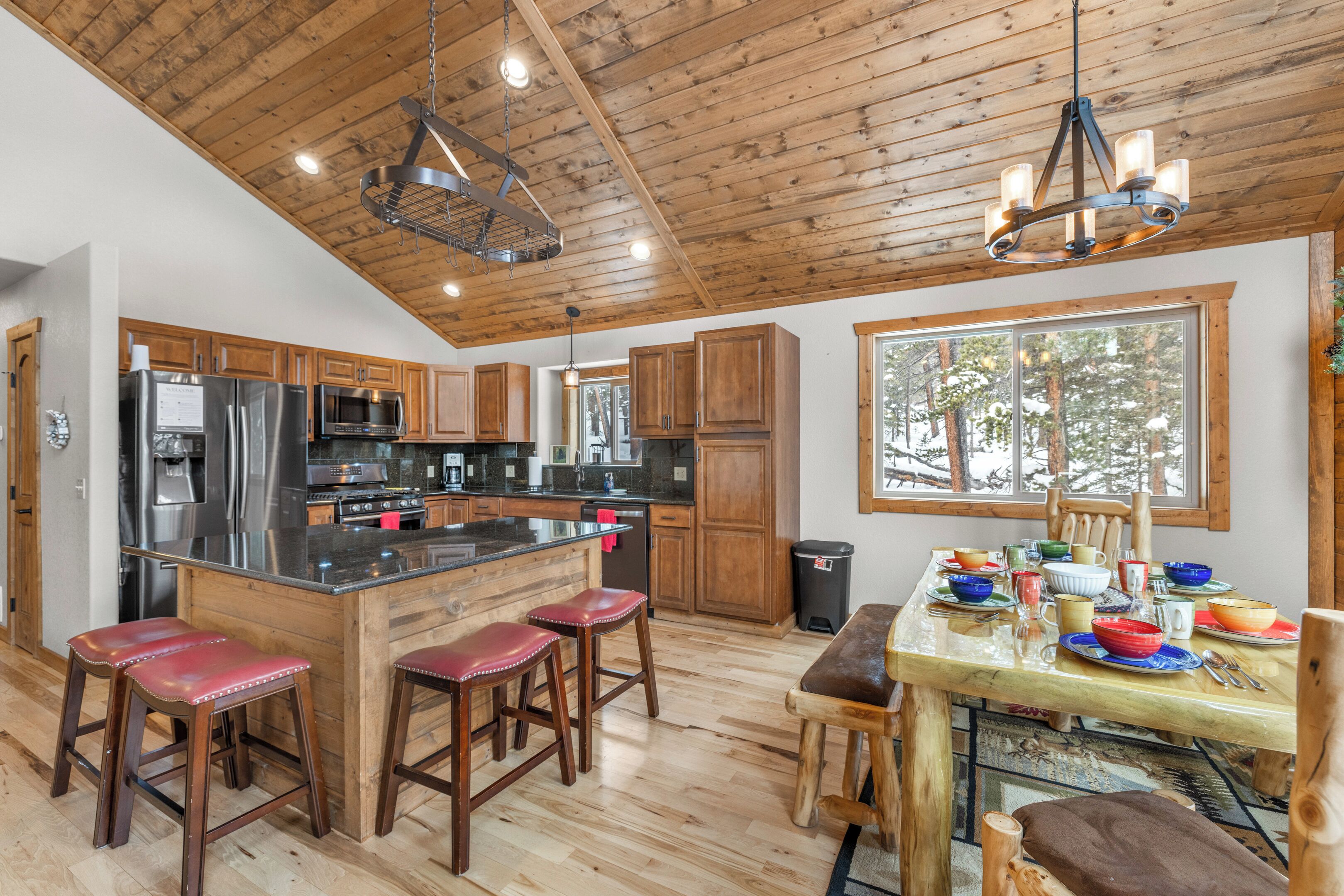 Une cuisine spacieuse avec des meubles en bois, un grand îlot central doté de tabourets, des appareils en acier inoxydable et une vue sur les arbres grâce à une grande fenêtre.
