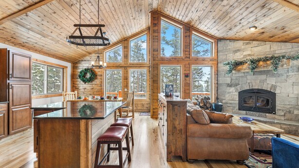 Interior espacioso de una cabaña con un amplio techo de madera, chimenea de piedra y múltiples ventanas con vistas al bosque circundante.