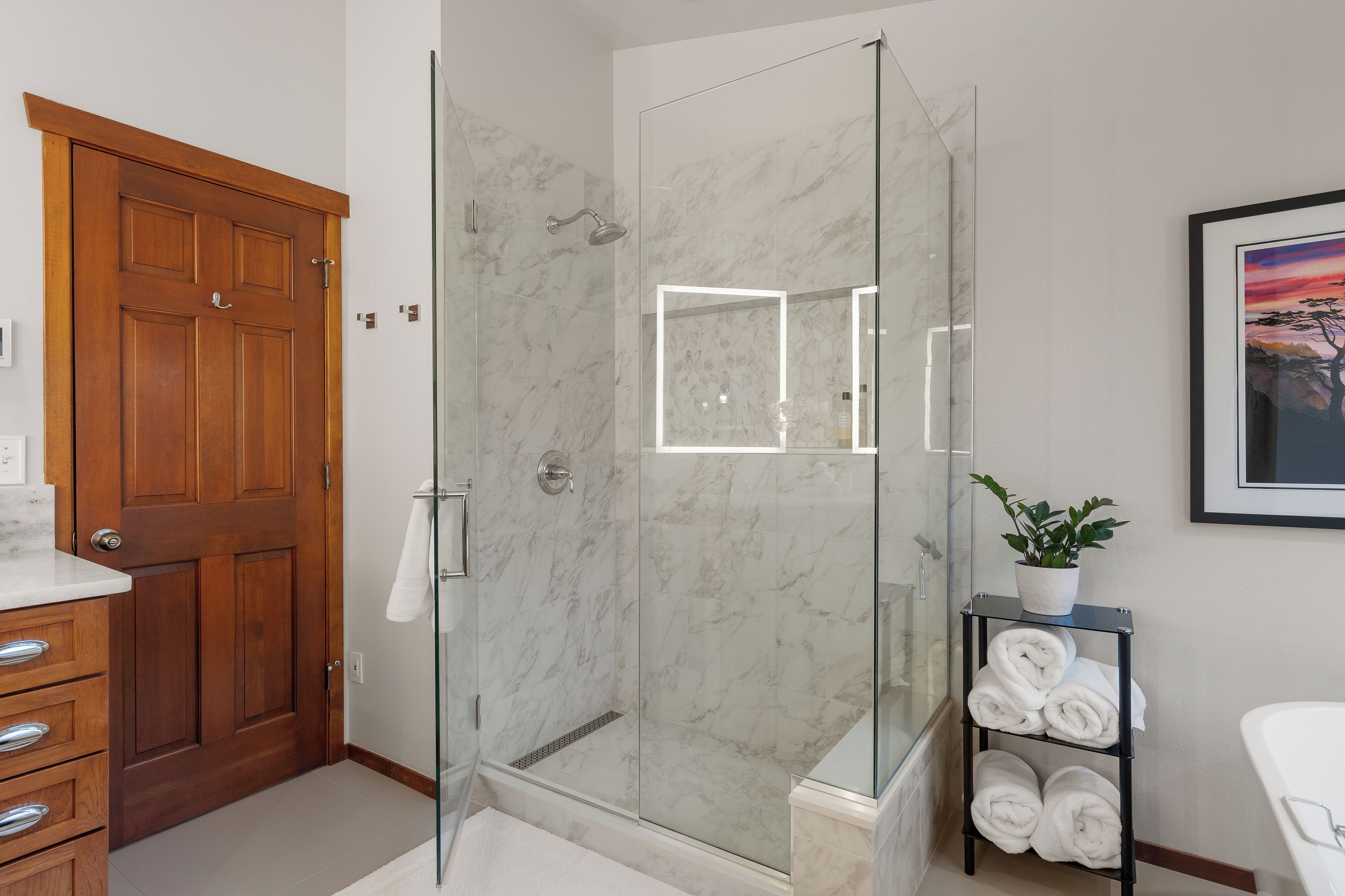 A modern bathroom with a glass-enclosed shower, marble walls, a wooden door, and a shelf with towels.