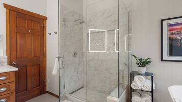 A modern bathroom with a glass-enclosed shower, marble walls, a wooden door, and a shelf with towels.