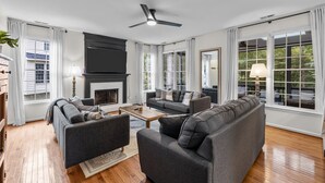 A modern living room with a fireplace, two sofas, a coffee table, and large windows with curtains. - Stunning 5 bedroom/3 bath home in Hope Valley Farm (Durham)