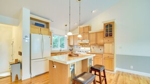 A modern kitchen with wooden cabinets, a white refrigerator, a central island with stools, and pendant lighting. - Spacious Lakefront New Auburn Home w/ Sunroom (New Auburn)