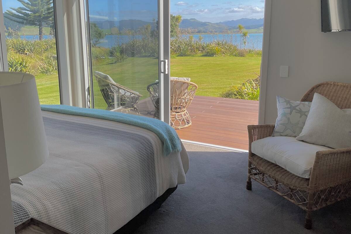 Ein Schlafzimmer mit Bett, einem Korbstuhl und Blick auf Garten und See.