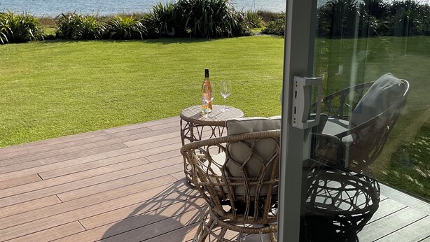 Una terraza con mobiliario de mimbre y una botella de vino, con vistas a un lago y montañas.