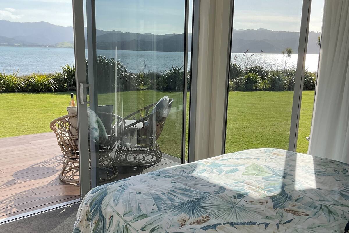 Ein Schlafzimmer mit einem großen Bett, Blick auf eine grüne Fläche und Wasser durch Glastüren sowie ein Korbstuhl an der Tür.