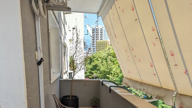Un balcón estrecho con una mesa, dos sillas, una planta en maceta y una ventana con toldo.