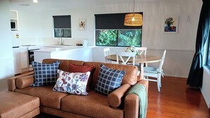 Ein Wohnzimmer mit braunem Sofa, einem Esstisch mit Stühlen, einer Küchenzeile und einem großen Fenster mit Grünpflanzen draußen.