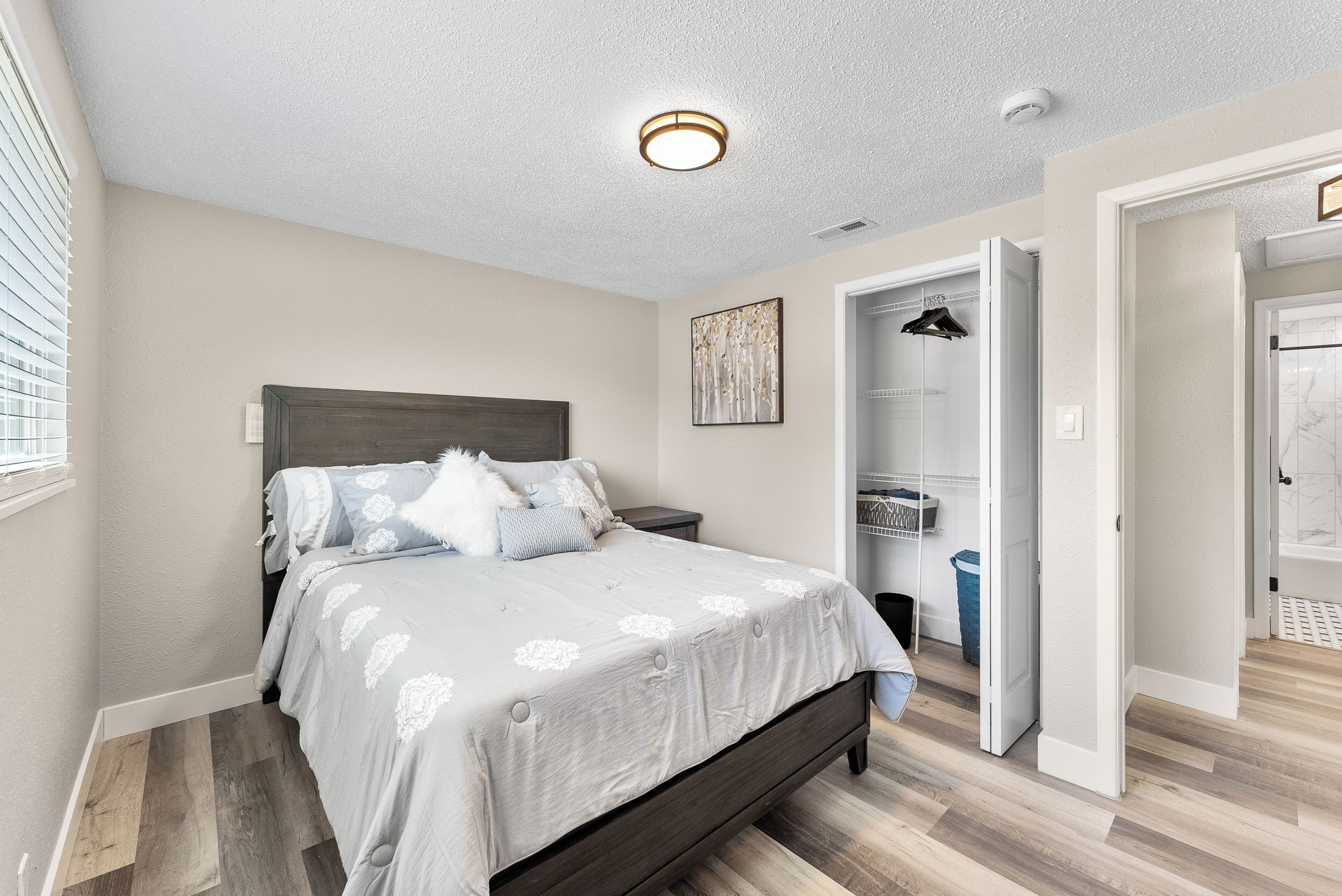 Une chambre à coucher comprenant un lit, une table de chevet, un luminaire fixé au mur, une porte donnant sur une salle de bain et un placard avec des vêtements suspendus.