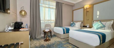 A hotel room with two beds, a desk with a coffee maker, a chair, a small table, and a large window with curtains.