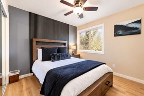 Une chambre à coucher avec un lit, un ventilateur de plafond, une fenêtre avec des stores et un tableau au mur.