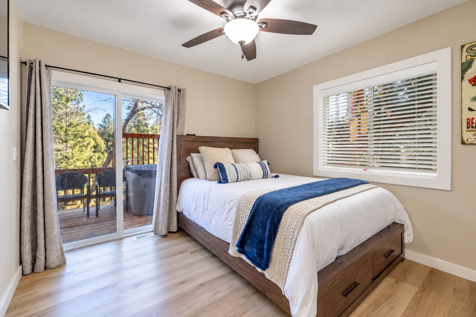 A bedroom with a bed, a ceiling fan, a window with blinds, and a balcony with a view of trees.