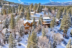 From above, you can see the cabin surrounded by pines, with snow-covered mountains and Big Bear Lake in the distance—giving you the full Big Bear setting from the moment you arrive. - Luxury 5BR Hot Tub · Fire Pit · GameRoom Sleep 11 (Big Bear Lake)