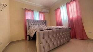 Een slaapkamer met een bed met een gestofte hoofdbord, roze gordijnen en een airconditioning aan de muur.