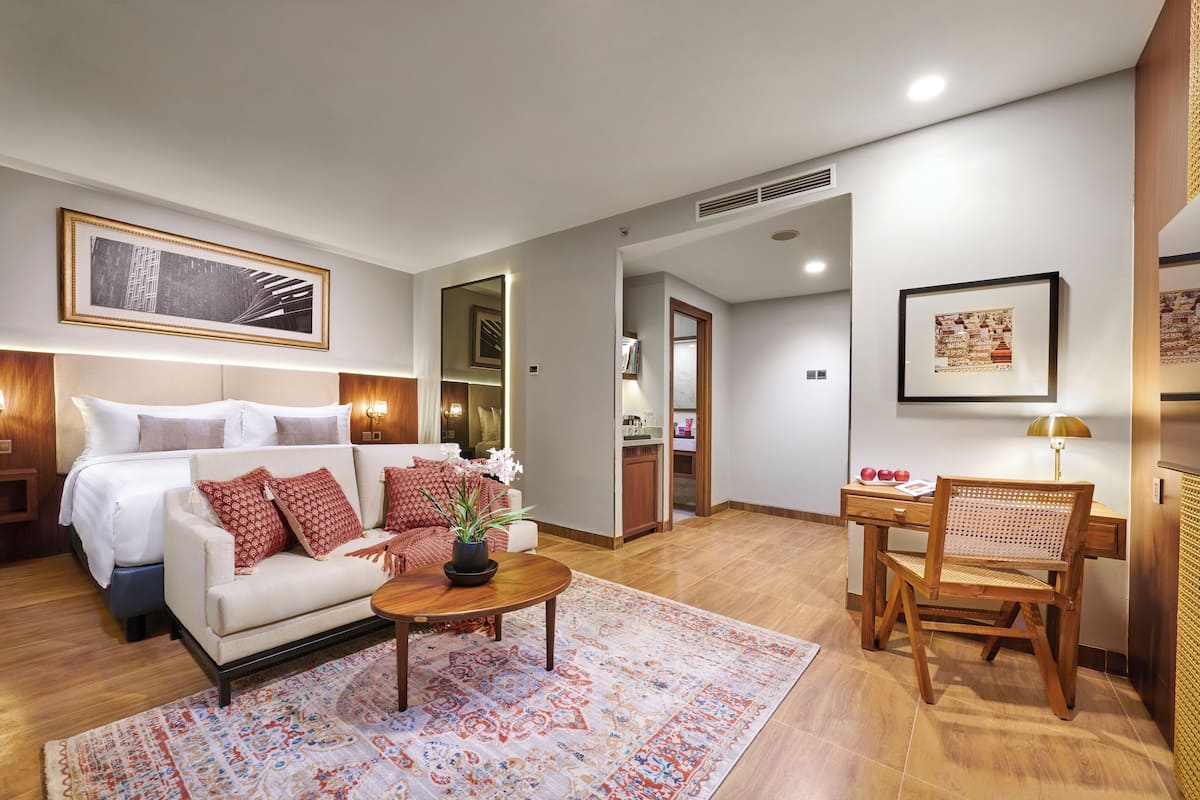 A modern hotel room with a bed, a sitting area with a coffee table and chairs, a desk with a lamp, and artwork on the walls.
