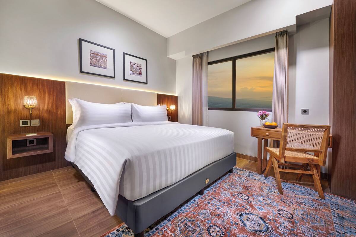 A hotel room with a large bed, a wooden nightstand with a lamp, a small table with a chair, and a large window with curtains.