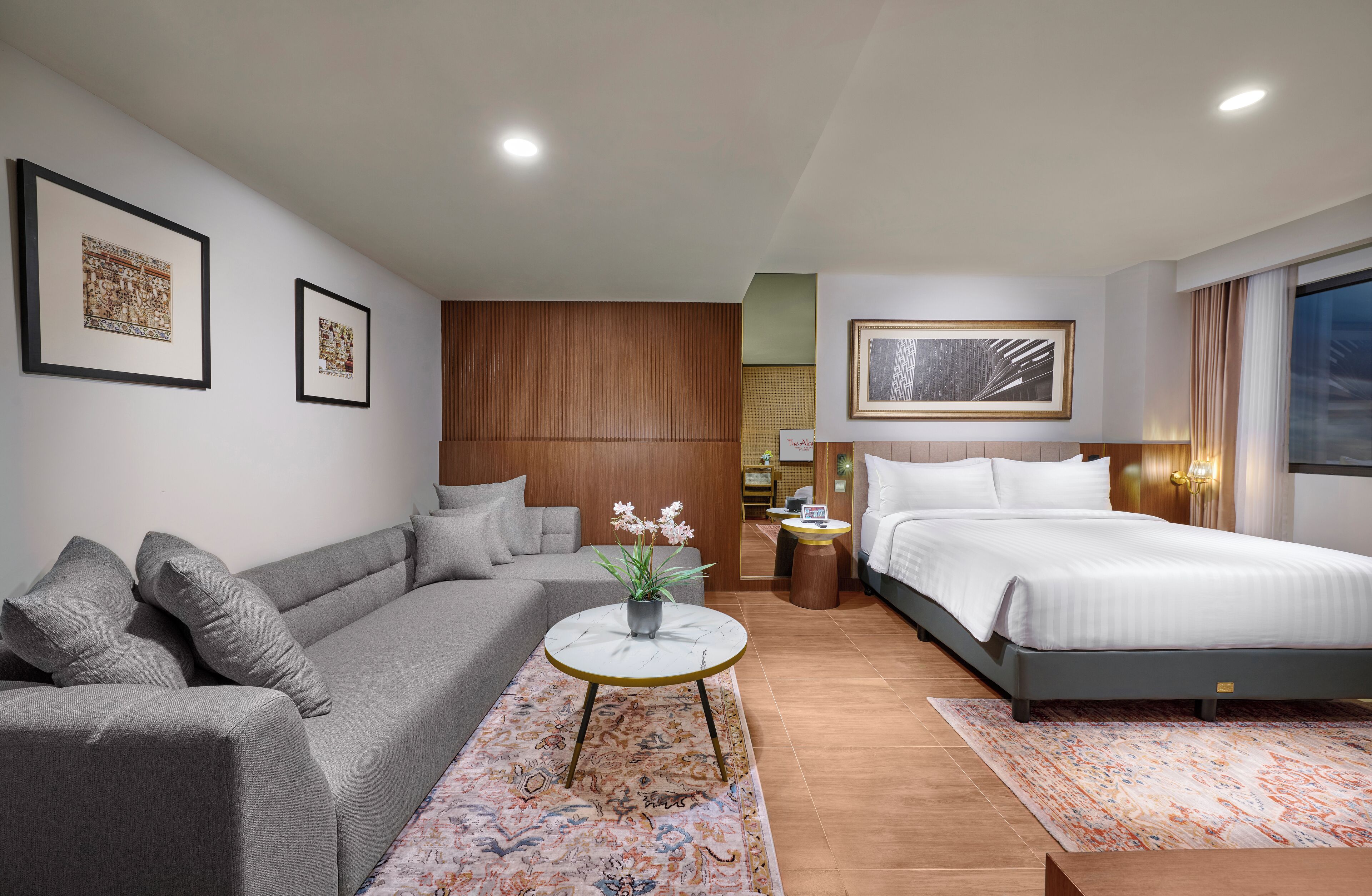 Una habitación de hotel moderna con un sofá gris, una cama, una mesita redonda con un florero y cuadros enmarcados en las paredes.