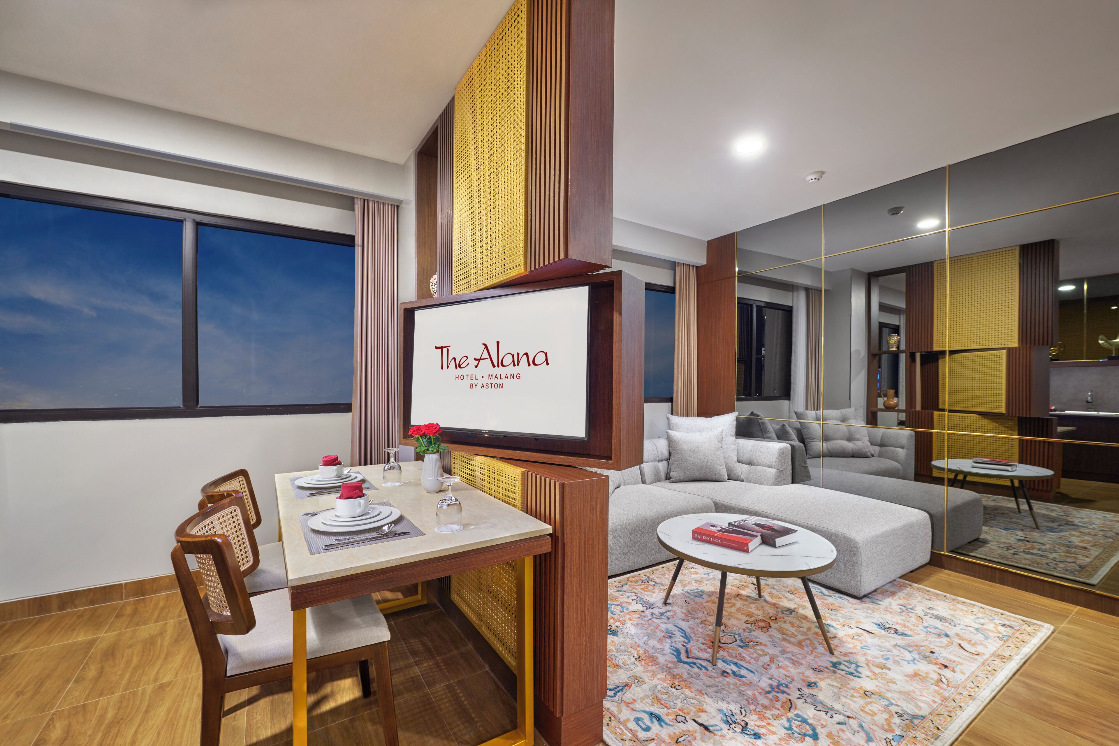 A modern hotel room with a dining area, a sofa, and a television displaying "The Alana Hotel - Malang by Aston".