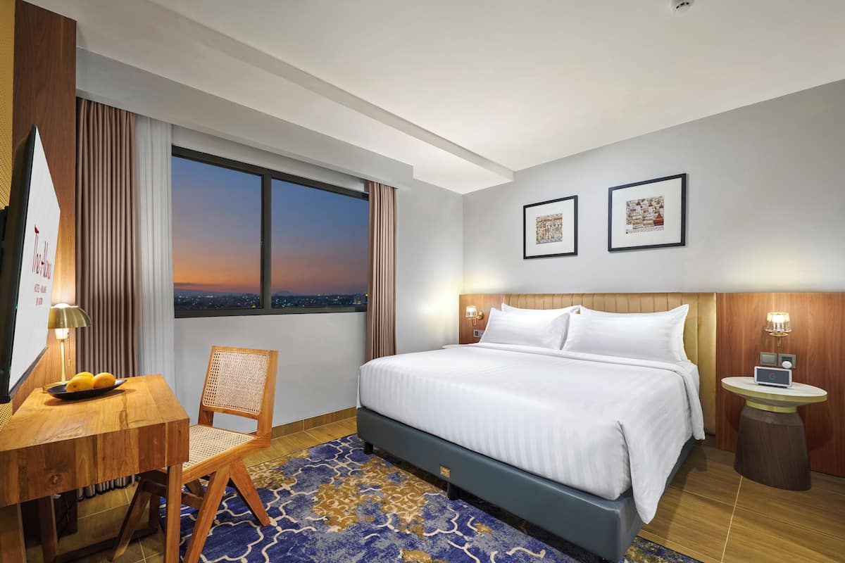 A hotel room with a large bed, a desk with a chair, a small table with a lamp, and two framed pictures on the wall.