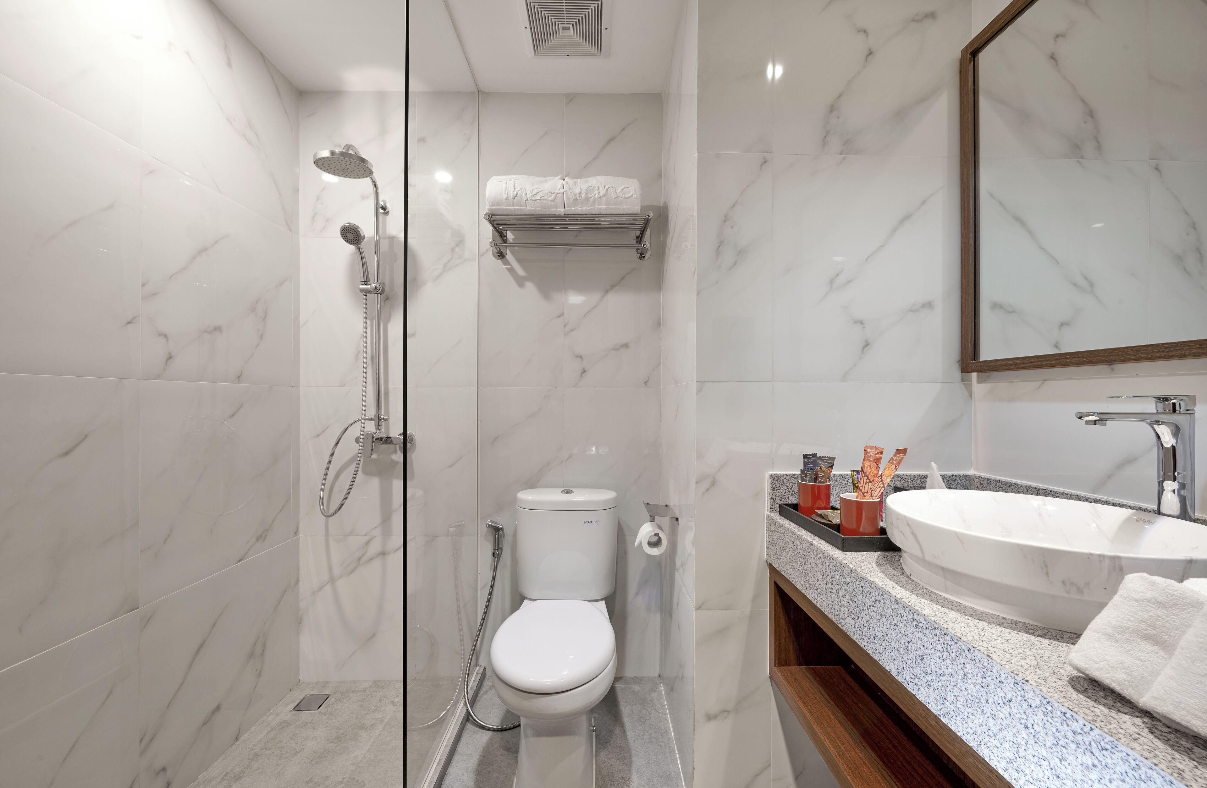 Uma casa de banho moderna com cabine de duche em vidro, sanita branca, parede revestida a mármore, lavatório com espelho e produtos de higiene pessoal no balcão.