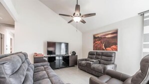 Een moderne woonkamer met een flatscreen televisie, leren banken, een plafondventilator en een groot schilderij aan de wand.
