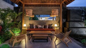 Ein Außenbereich mit einer hölzernen Bank, einem Sofa und zwei Stühlen. Auf beiden Seiten des Sofas stehen jeweils eine Tischlampe.