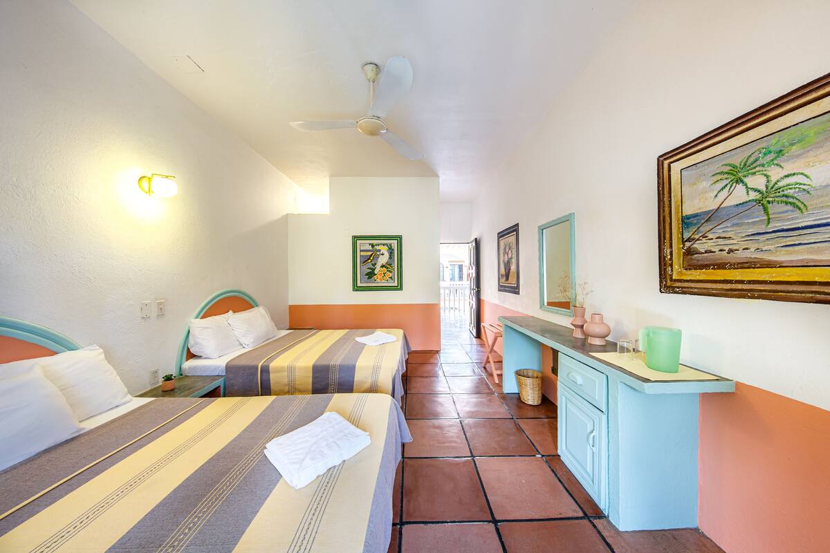 Habitación de hotel con dos camas, un ventilador de techo, un escritorio con un florero y un cuadro de una playa.