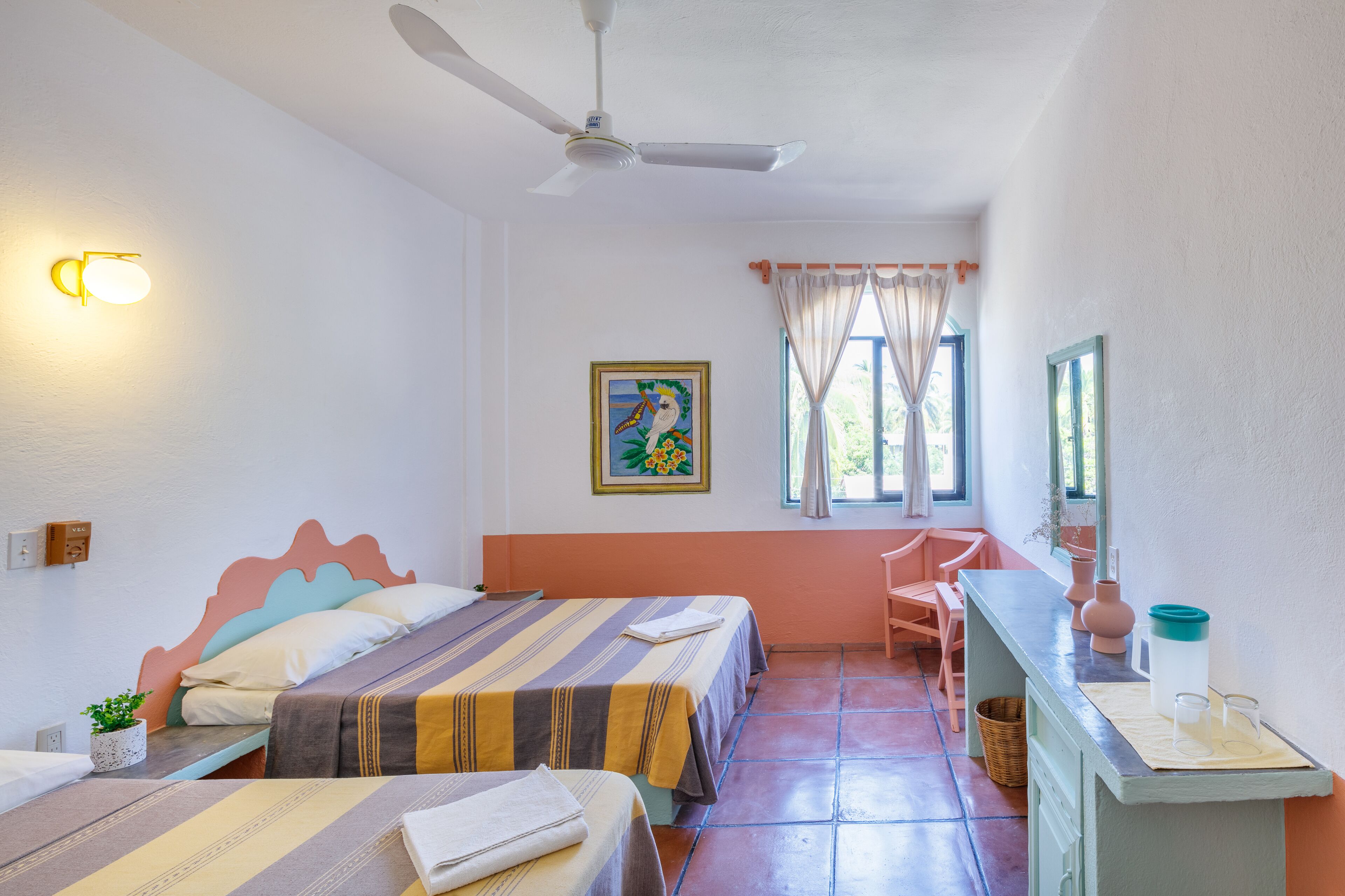 A room with two beds, a desk, a chair, a ceiling fan, and a window with curtains.